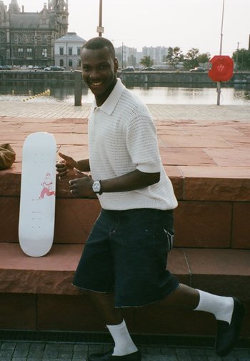 Witte skateboard met een rode graphic van een rennend figuur, geplaatst op een roodachtige stenen oppervlakte. Persoon in een wit gebreid polo en denimshorts poseert in de buurt.