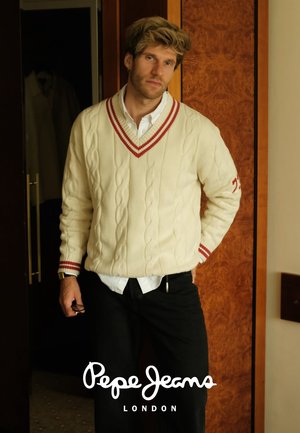 Suéter de punto trenzado color crema con cuello en V y puños a rayas rojas, llevado sobre una camisa blanca, combinado con pantalones oscuros, de pie en el interior.