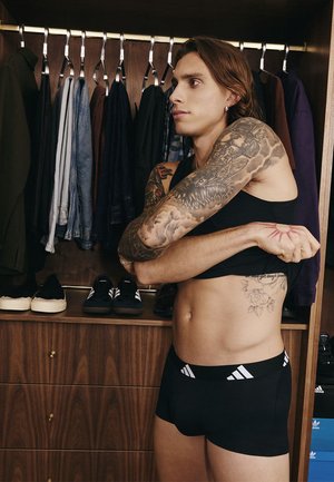 Persona con tatuajes que lleva ropa deportiva negra estira el brazo en un armario de madera lleno de ropa colgada y zapatos.