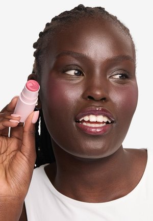 Donna con pelle scura che tiene vicino alla guancia un bastoncino di blush crema rosa, indossa una maglietta bianca, sorride e guarda di lato.