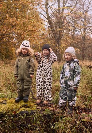 Drie kinderen staan op een stam in een grasrijke omgeving. Ze dragen kleurrijke geïsoleerde overalls: olijfgroen, luipaardprint en een camouflagepatroon.