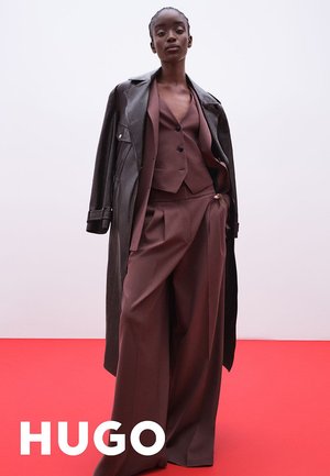 Costume bordeaux avec un pantalon à jambes larges et un gilet ajusté sous un manteau en cuir noir, sur fond rouge.