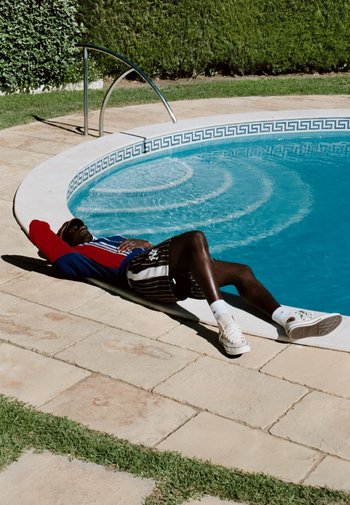 A person lounges beside a circular pool, wearing a red and blue jacket, striped shorts, and white sneakers. The area features stone tiles and greenery.