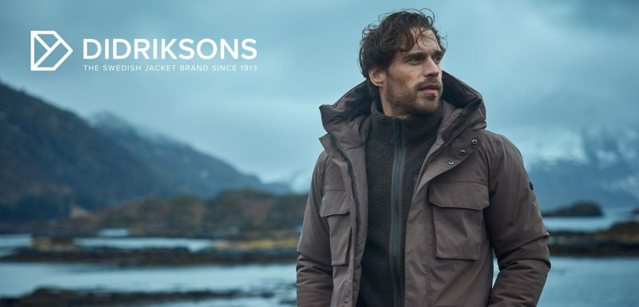 Braune Kapuzenjacke mit einem Reißverschluss vorne, großen Taschen und einem hohen Kragen, getragen über einem dunkel strukturierten Pullover, vor einem maritimen Hintergrund.