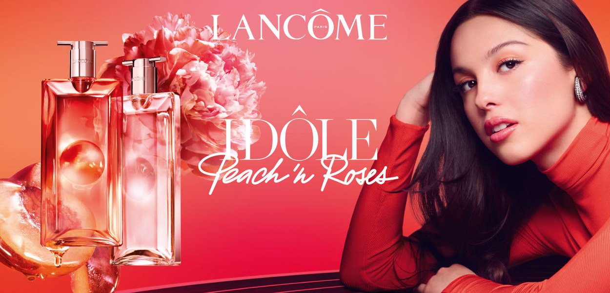 Jeune femme vêtue d'un haut rouge à côté de flacons de parfum Lancôme Idôle aux tons pêche et rose, avec des fleurs roses sur un fond dégradé pêche.