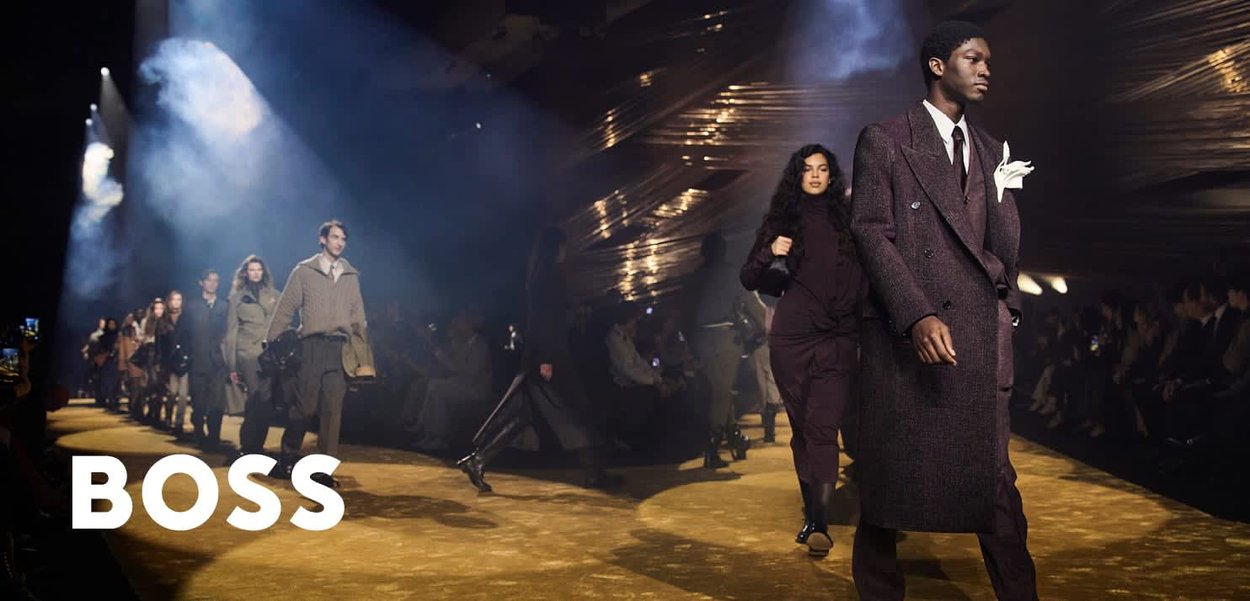 Modele prezentujące stylowe jesienno-zimowe stroje idą po wybiegu podczas przyciemnionego pokazu mody w pomieszczeniu, z widownią na miejscach siedzących.