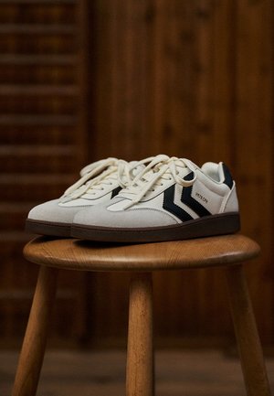 White suede sneakers with black accents, round toe, padded collar, and flat laces on a wooden stool. Brown rubber outsole.