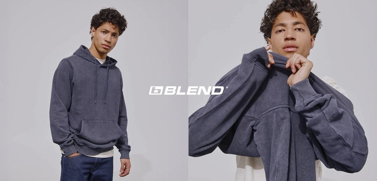 Jonge man met krullend haar die een donkergrijze hoodie over een licht shirt aantrekt tegen een effen achtergrond, met het logo "BLEND" eroverheen.
