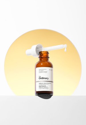Botella de vidrio marrón de suero facial The Ordinary con gotero blanco dispensando una gota, fondo circular amarillo.