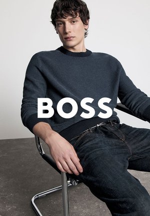 Gestickter navyfarbener Pullover mit gerippten Bündchen, kombiniert mit dunklen Jeans. Model sitzt auf einem Chromstuhl vor einem minimalistischen Hintergrund.