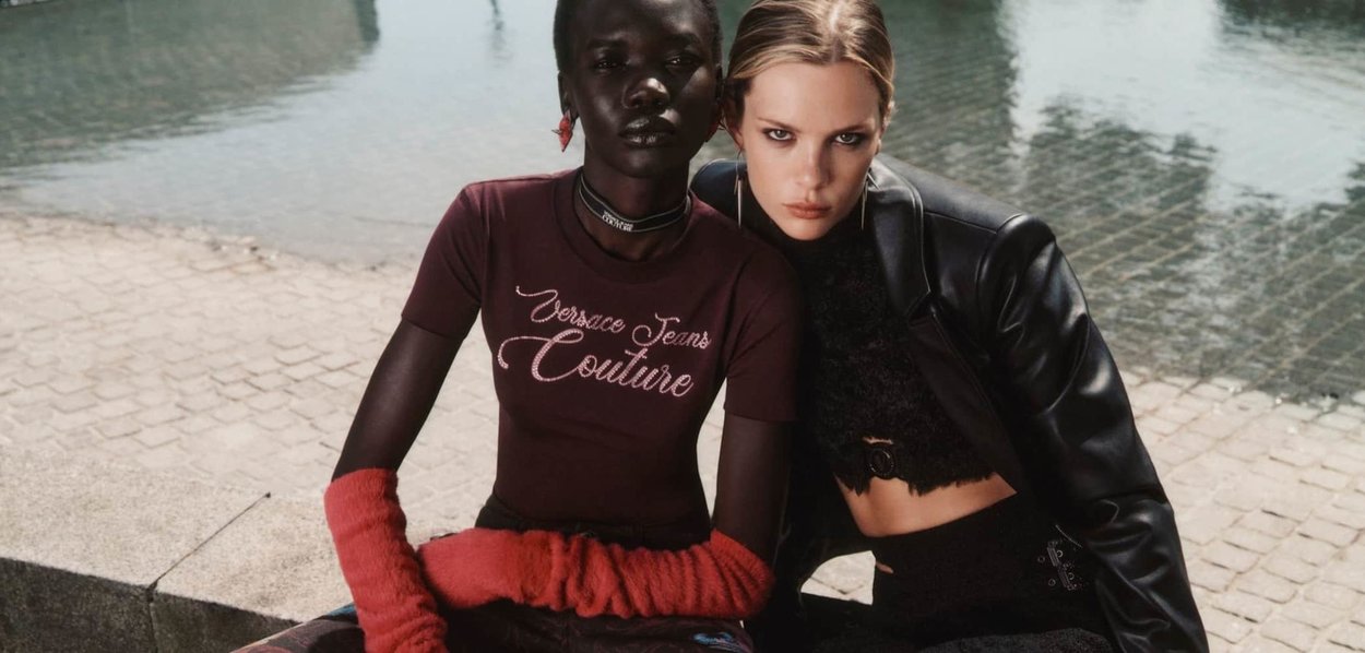 Due modelli siedono insieme. Uno indossa un top bordeaux con "Versace Jeans Couture" in bianco, abbinato a guanti pelosi rossi. L'altro indossa una giacca di pelle nera.