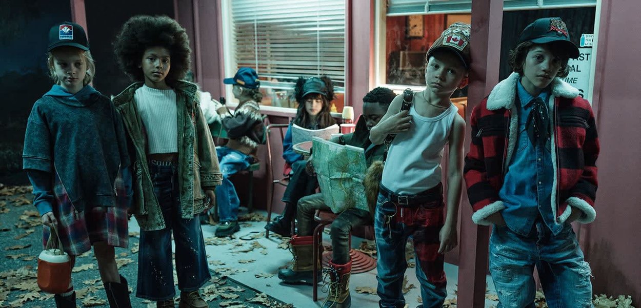 Zeven kinderen, gekleed in herfstkleding, staan en zitten voor een diner, sommigen houden kaarten vast en dragen hoeden, met herfstbladeren op de grond.