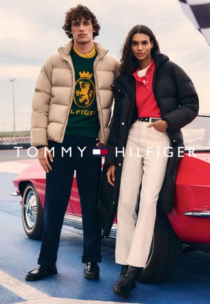 Abrigo acolchado beige claro, suéter verde con logo, chaqueta negra, polo rojo, pantalones blancos de cintura alta, botas negras, posando junto a un coche vintage.