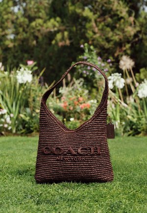 Borsa intrecciata marrone con una tracolla singola e logo "Coach New York", posata sull’erba con fiori da giardino e vegetazione sfocati sullo sfondo.