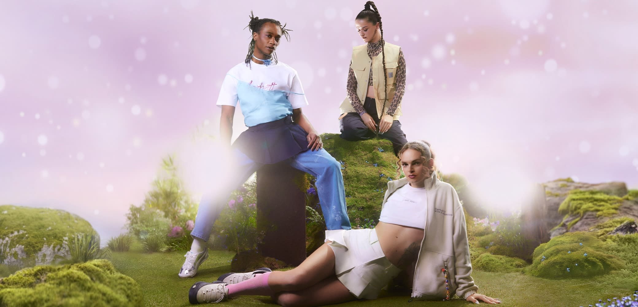 Trīs modeļi pozē uz zālaina seguma. Apģērbu komplektā ietilpst slāņots zils un balts krekls, bēša veste un balta jakas ar akcentiem.