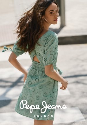 Mujer joven con el pelo largo que lleva un vestido verde menta con ojales, mangas abullonadas y una apertura en la espalda, caminando al aire libre por una calle soleada.