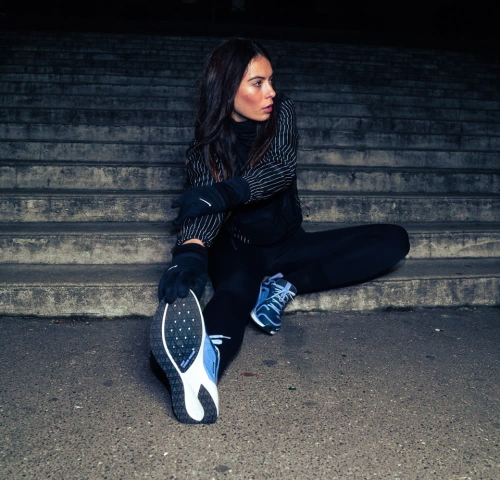 Athletic footwear features a blue and white design with textured soles. The model wears a black fitted outfit with distinct striped accents and gloves.