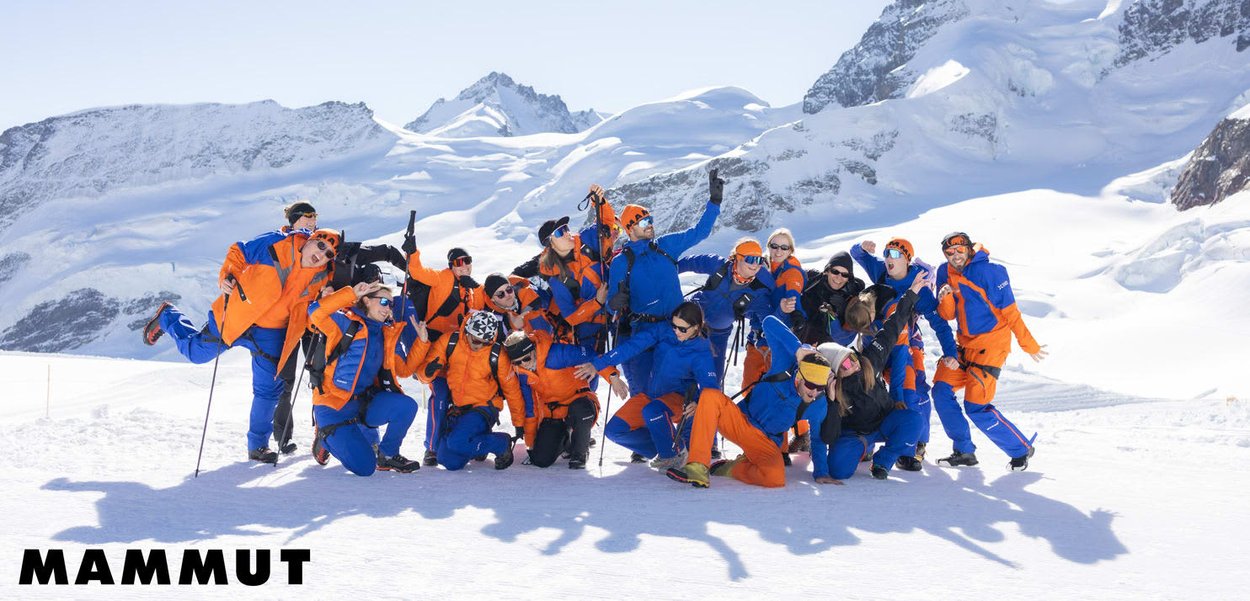 Gruppe von 19 Personen in orangefarbener und blauer Winterbekleidung, die spielerisch im Schnee posieren, mit schneebedeckten Bergen im Hintergrund und klarem Himmel.