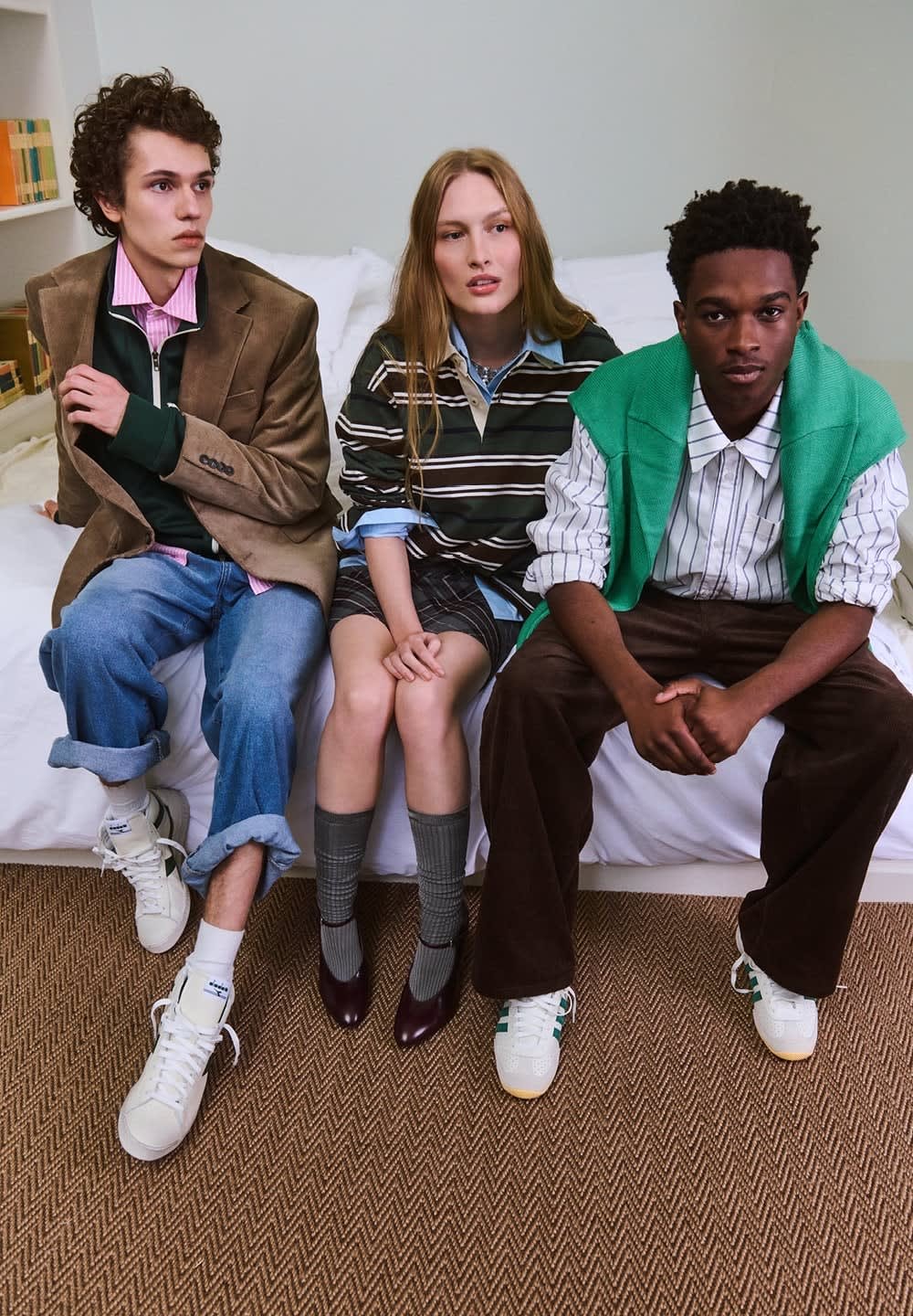 Kolme henkilöä sängyllä: vasemmalla ruskea bleiseri ja vihreä paita, keskellä raidallinen yläosa ja ruudullinen hame, oikealla vihreä liivi raidallisen paidan päällä.