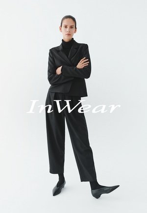 Schwarzer, maßgeschneiderter Blazer und hoch taillierte Hose aus glattem Stoff, kombiniert mit spitzen schwarzen Ballerinas und einem eng anliegenden schwarzen Rollkragenpullover.