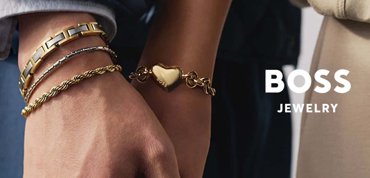 Nahaufnahme von zwei Handgelenken, die goldene Armbänder tragen, darunter ein herzförmiges Charms-Armband mit der Gravur "BOSS", vor einem dunklen und hellen Hintergrund.