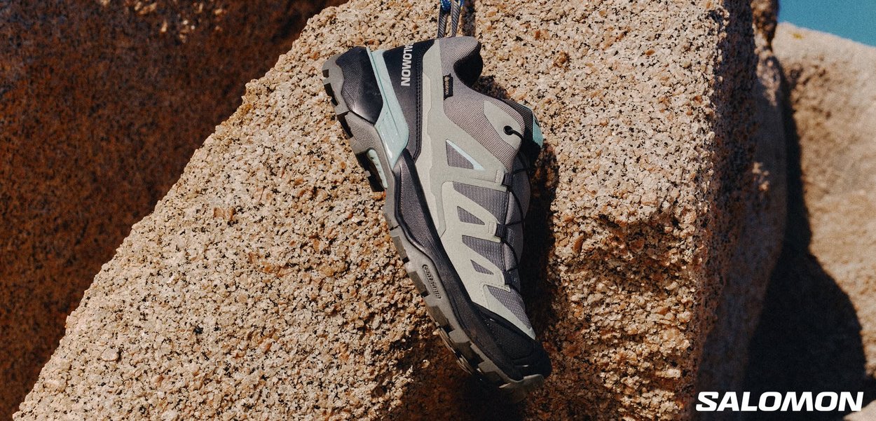 Zapatilla deportiva gris y negra con acentos turquesa, que presenta una suela texturizada, sistema de cordones y un acabado gomoso, descansando sobre una superficie rocosa.