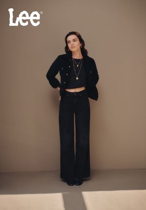 Schwarze Jeansjacke mit Messingknöpfen, kombiniert mit einem schwarzen Cropped-Top und hoch taillierten, weit geschnittenen schwarzen Jeans, accessorized mit geschichteten Halsketten.
