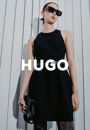 Vestido negro sin mangas con cuerpo ajustado y falda acampanada, adornado con un cinturón plateado. La modelo sostiene un pequeño bolso negro.