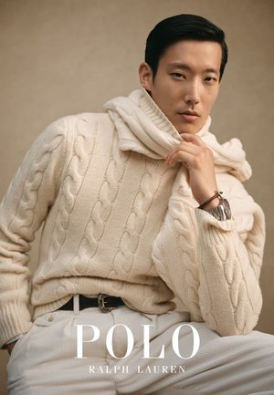 Kremfarget kabelstrikket genser med høy krage, iført lyse bukser. Merkbare detaljer inkluderer et ur og en avslappet positur.