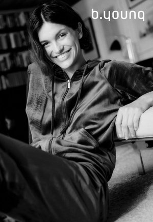 Schwarz-weißes Bild einer Frau in einem Velours-Trainingsanzug mit Reißverschluss und Kapuze, mit glatter Textur und seitlichen Taschen, entspannt sitzend.