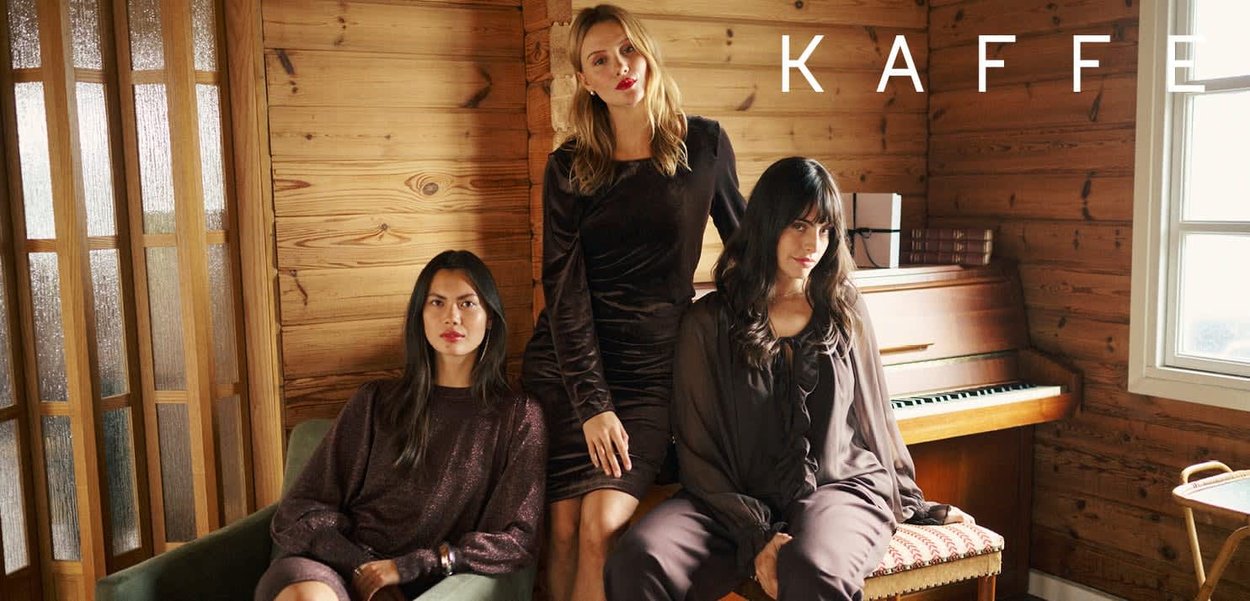 Tre kvinder iført tekstureret mørkt tøj poserer indendørs foran en trævæg. Et klaver og bøger er synlige i baggrunden.