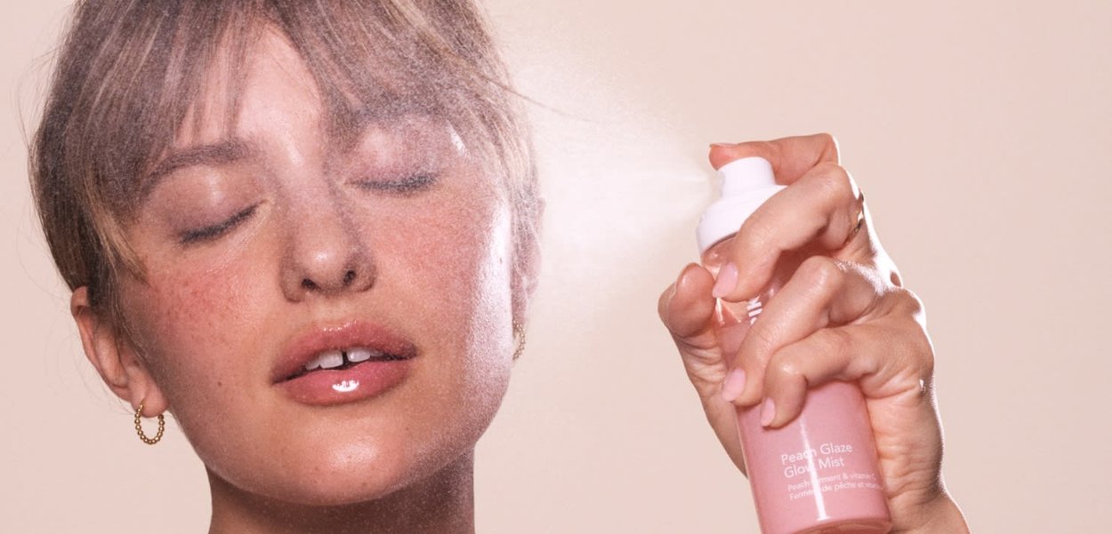 Pink spray bottle labeled "Peach Glaze Glow Mist," being misted on the face. Smooth texture, minimalistic design, and soft lighting.