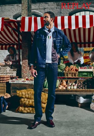 Jeansjakke med trykknapper, hvit polo med italiensk flagg, mørke jeans og brune sko, satt mot en marked bakgrunn med frukt og grønnsaker.