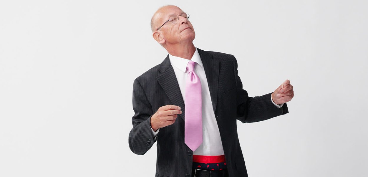 Mann in einem gestreiften Anzug mit einem weißen Hemd und einer pinken Krawatte, der einen roten Gürtel über blauen Hosen mit einem Muster trägt. Weißer Hintergrund.