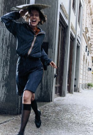 Chaqueta impermeable con rayas de colores y falda a juego, usada con calcetas hasta la rodilla. Corriendo por una calle empedrada mojada, sosteniendo un paraguas.