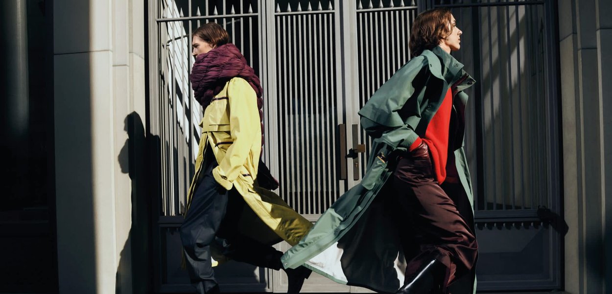Deux mannequins marchent rapidement, l'un portant un trench jaune avec une grande écharpe bordeaux et l'autre un trench vert sur un haut rouge, un pantalon marron et des bottes.