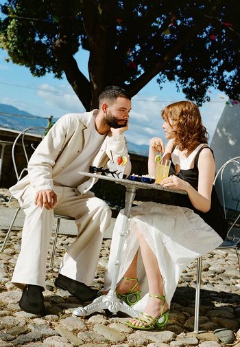 Due persone sedute a un tavolo rotondo. L'uomo indossa un abito beige, mentre la donna indossa un vestito nero con una gonna in pizzo bianco e sandali verdi con cinturini.