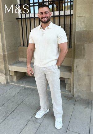 Man glimlacht, draagt een wit gebreide polo met korte mouwen, lichtbeige broek en witte sneakers, staand voor een stenen muur met een raam met tralies.