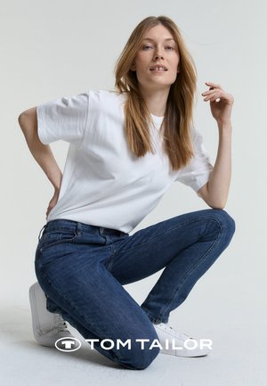Bianco t-shirt in cotone, jeans in denim blu e sneakers bianchi. I capelli del modello sono lisci e a strati. La posa è in ginocchio con una mano sui fianchi.
