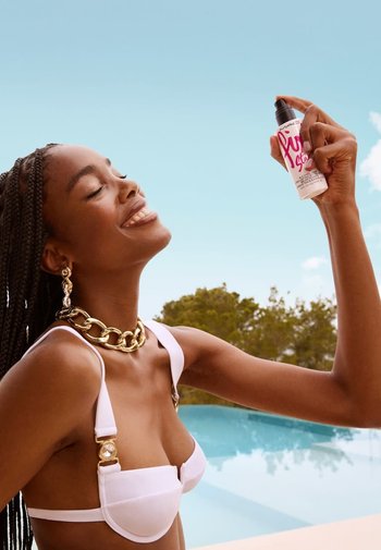 Bottiglia spray bianca con testo rosa tenuta da una persona che indossa un bikini bianco e gioielli d'oro massiccio. Sfondo con piscina e alberi.