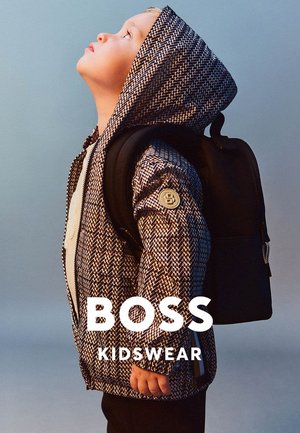 Enfant portant une veste à capuche à motifs et un sac à dos noir, regardant vers le haut devant un fond uni, avec le texte "BOSS KIDSWEAR".