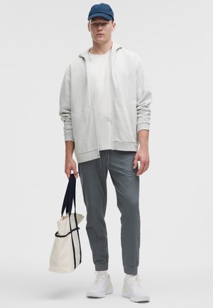 Vaaleanharmaa vetoketjullinen huppari, jossa etutaskut, valkoinen t-paita, slim-fit-harmaat housut ja valkoiset tennarit. Pitelee beigeä kangaskassia, jossa siniset yksityiskohdat.