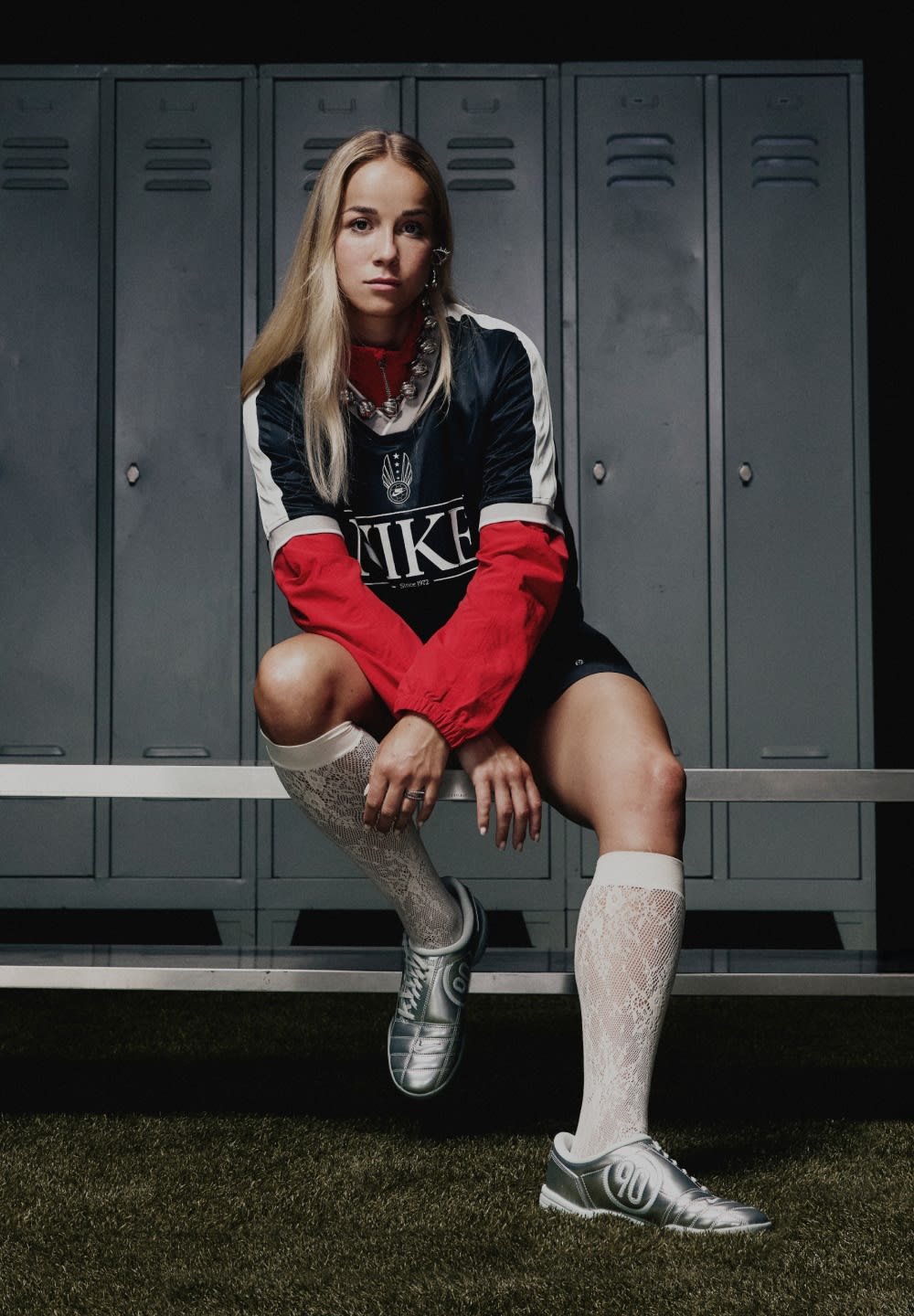 Athlete in a black and red Nike jersey, silver shoes, and white lace socks sits on a bench in front of gray metal lockers.