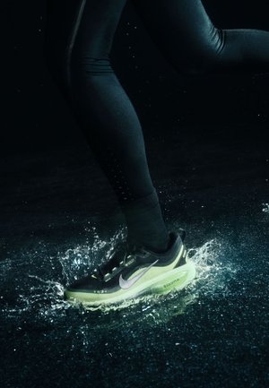 Schwarzer Laufschuh mit grüner Sohle und lila Akzenten, der durch Wasser spritzt. Getragen mit schwarzen Leggings, die eine längsgerichtete Textur aufweisen.