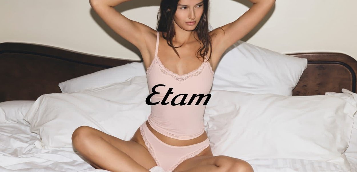 Giovane donna in lingerie rosa chiaro con rifiniture in pizzo, seduta a gambe incrociate su un letto bianco, con le braccia tese dietro la testa, sfondo neutro.