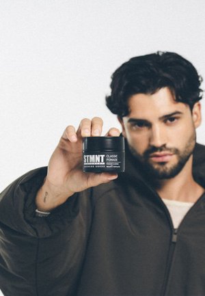 STMT Classic Pomade in een zwarte verpakking met witte tekst. Het product wordt vastgehouden door een persoon die een bruine jas draagt en heeft een matte afwerking.