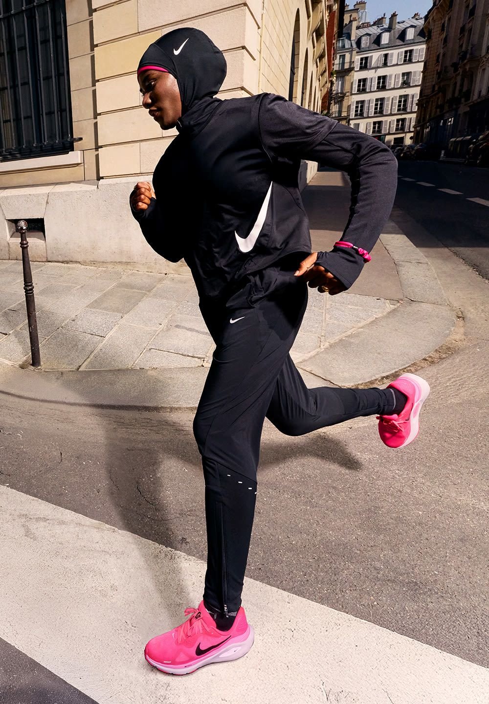 Completo sportivo nero con cappuccio, caratterizzato da un logo Nike bianco. Scarpe da corsa rosa e tessuto strutturato. Ambientazione urbana con pavimentazione e edifici.