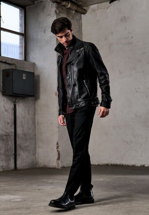 Giacca nera in pelle con colletto a revers, abbinata a una maglia color bordeaux e pantaloni neri. La modella posa in un ambiente industriale, mostrando un design elegante.