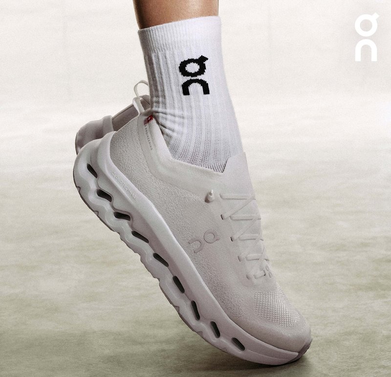 Weißer Sportschuh mit einem strukturierten Mesh-Obermaterial, ausgeprägtem Dämpfungsmittelsohle und einem Logo an der Seite. Tragen mit einer weißen Socke mit schwarzem Logo.