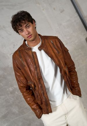 Brown leather bomber jacket with a high collar, zippered front, and side pockets, worn over a white shirt and light pants on a neutral background.